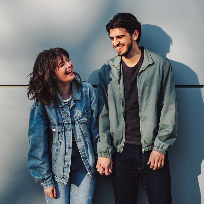 Smiling girlfriend holding hand of her boyfriend while laughing and standing together in front of the gray wall at the city street.
